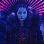 Ana de Armas as the character Rooney in Ballerina, wearing a fur-collared coat and a sequined top, with a serious expression, standing in a vibrant, neon-lit tunnel with pink and blue stripes.