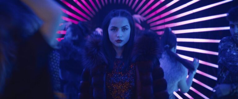Ana de Armas as the character Rooney in Ballerina, wearing a fur-collared coat and a sequined top, with a serious expression, standing in a vibrant, neon-lit tunnel with pink and blue stripes.