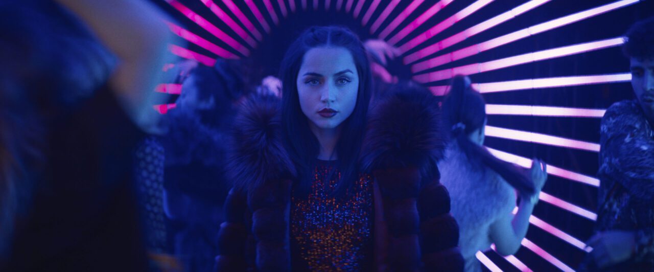 Ana de Armas as the character Rooney in Ballerina, wearing a fur-collared coat and a sequined top, with a serious expression, standing in a vibrant, neon-lit tunnel with pink and blue stripes.