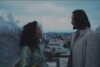 Keanu Reeves, with large white wings attached to his back, looking intently at a woman with dark hair, on a rooftop or balcony overlooking a cityscape at dusk, from the movie Good Fortune.