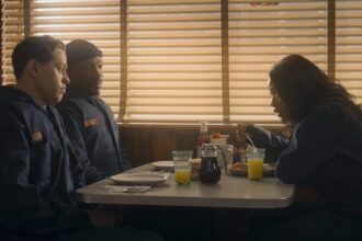 Three individuals, two men in what appear to be work uniforms and a woman eating, seated in a diner booth with venetian blinds on the windows, from the movie The Pickup.