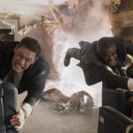 John Cena and Idris Elba, dressed in suits, struggle to hold onto seats in a rapidly disintegrating airplane cabin filled with smoke and debris, their faces strained.