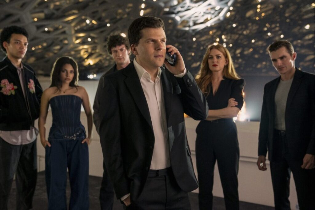 J. Daniel Atlas (Jesse Eisenberg) on a phone, surrounded by several other cast members, including Isla Fisher as Henley, and others, in what appears to be a large, modern, possibly event space, in Now You See Me, Now You Don't.