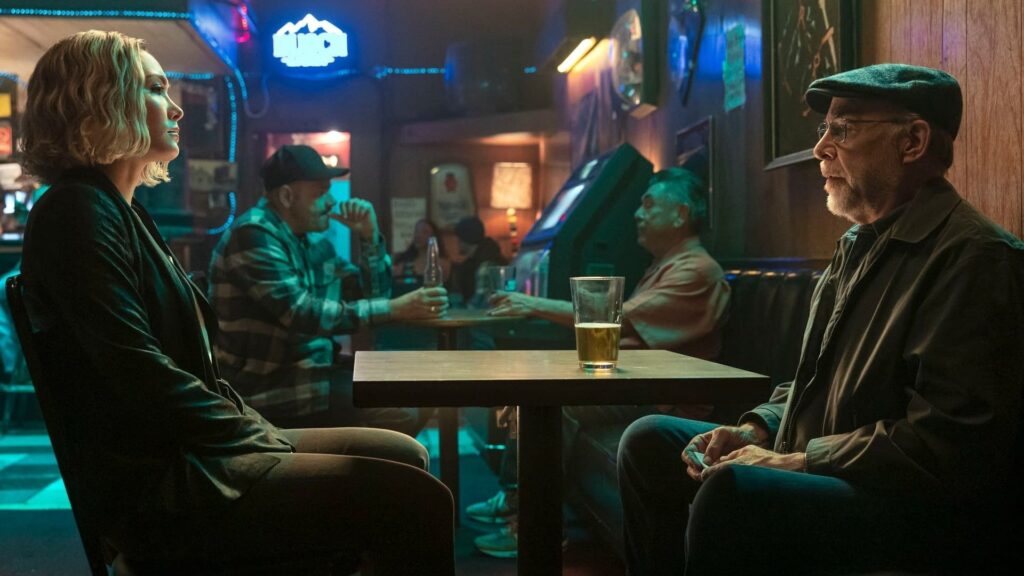 Movie still from The Accountant 2 - Raymond King (J.K. Simmons) in a diner booth, sitting across from Anaïs, in a dimly lit, blue-hued setting.